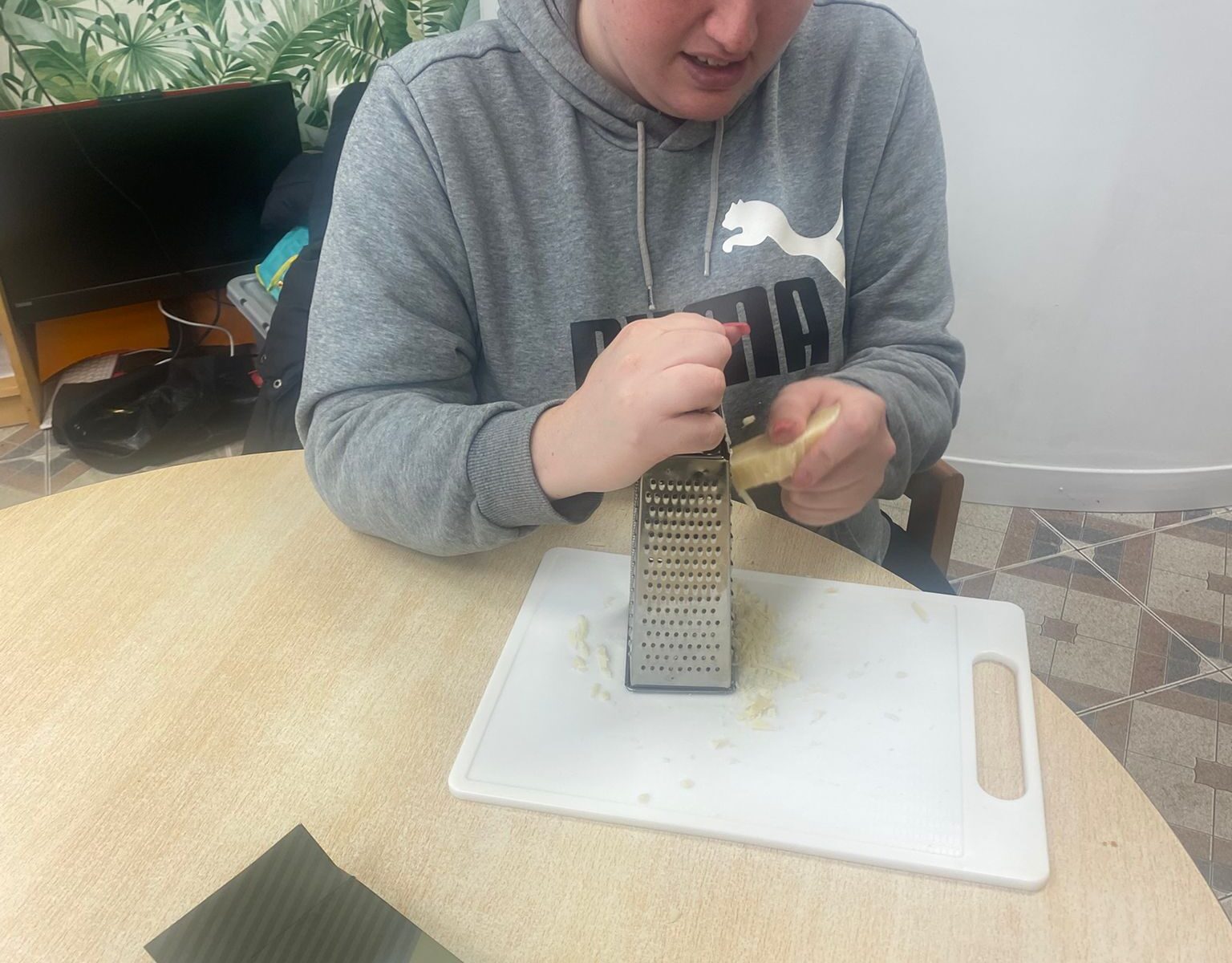 A woman in a grey Puma sweatshirt grates cheese on a white cutting board in preparation for cooking. A box of lasagne sheets and a baking tray sit on the table beside her.