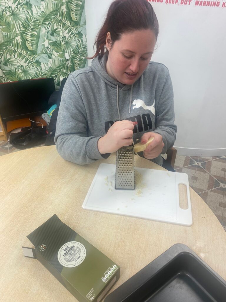 A woman in a grey Puma sweatshirt grates cheese on a white cutting board in preparation for cooking. A box of lasagne sheets and a baking tray sit on the table beside her.