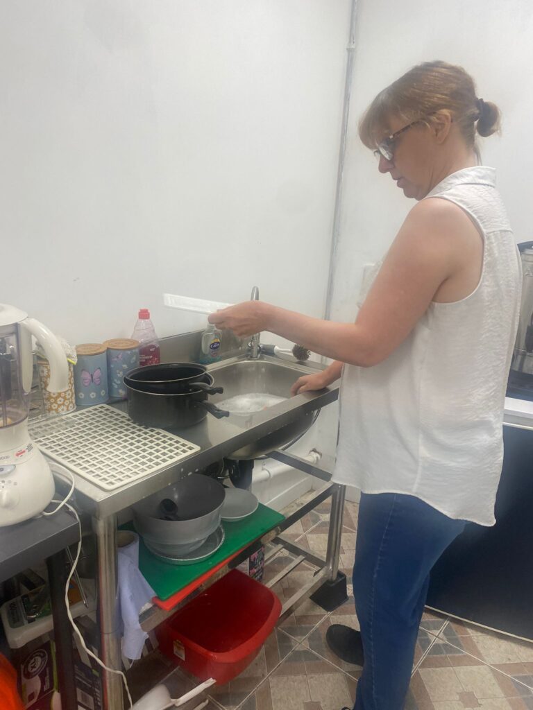 A woman in a white sleeveless top stands at a sink, washing dishes. A variety of kitchen tools and cleaning supplies are visible on the counter around her.