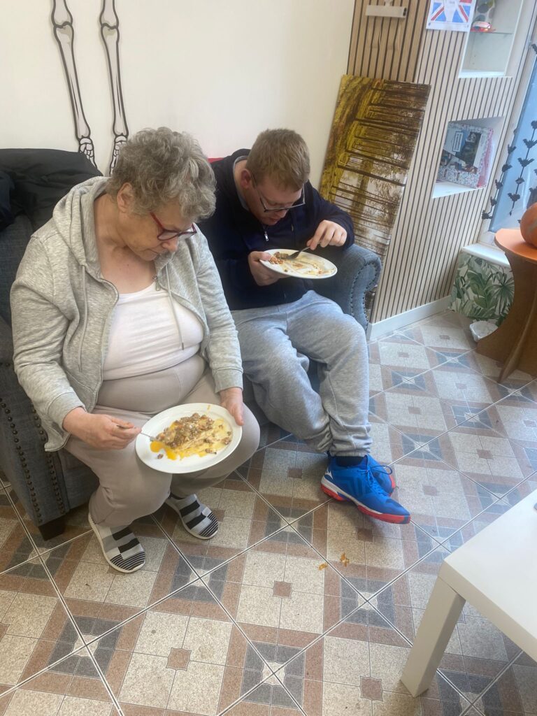 Two people, an elderly woman and a younger man, sit on a grey couch enjoying plates of food. They appear to be focused on their meals, with empty plates nearly visible.