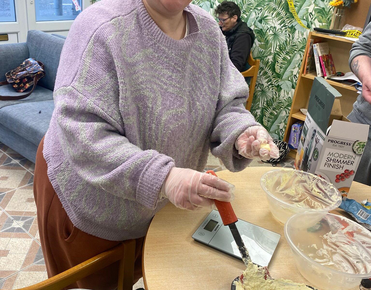 A smiling woman in a purple sweater spreads cheesecake filling into a pan. She wears gloves, and kitchen tools and ingredients are arranged on the table around her.