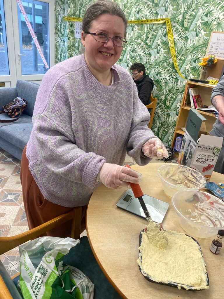 A smiling woman in a purple sweater spreads cheesecake filling into a pan. She wears gloves, and kitchen tools and ingredients are arranged on the table around her.