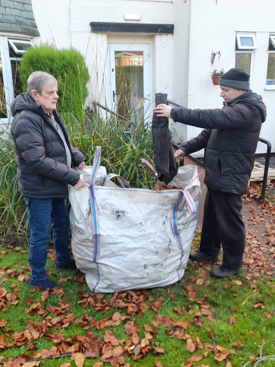 Two individuals, bundled up in warm clothing, are seen working together to fill a large garden bag with fallen autumn leaves. They stand on a grassy area, with leaves scattered around, in front of a light-colored building.