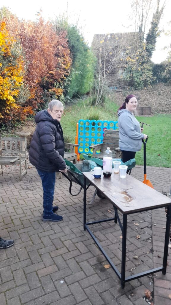 A man in a dark jacket pulls a green garden trolley, while a woman in a grey hoodie holds a shovel, standing in a paved area. There are some mugs and a bottle on a nearby table, with autumn-colored bushes in the background.