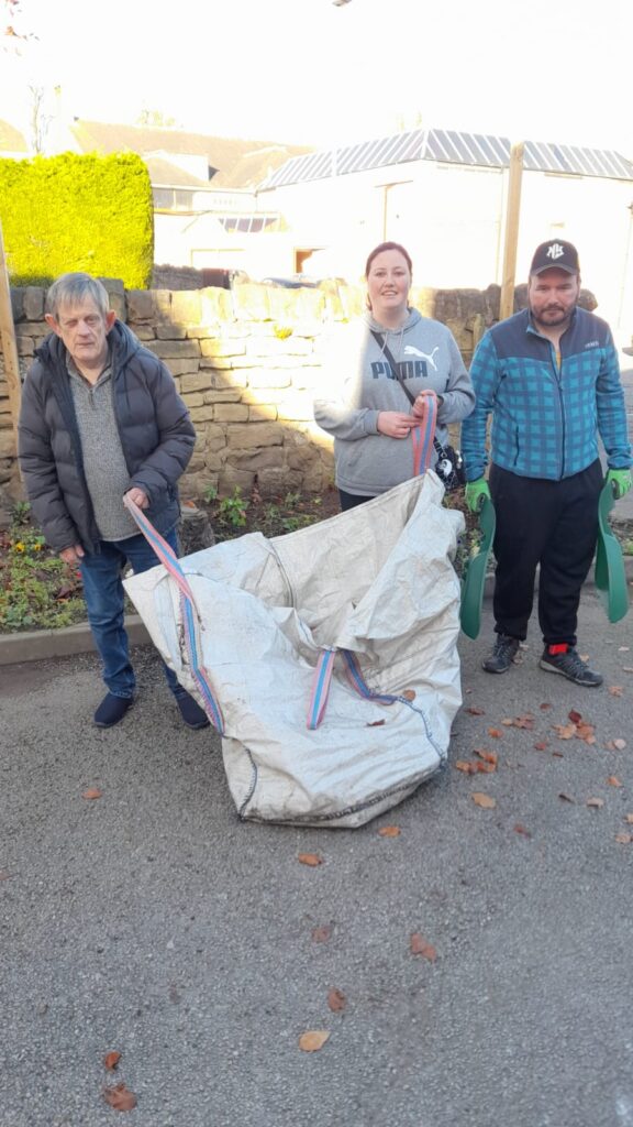 Three people posing for a photo outdoors, holding a large, partially filled garden bag. They are standing on a cleared driveway with a stone wall and greenery behind them, dressed in casual outdoor clothing for cooler weather.