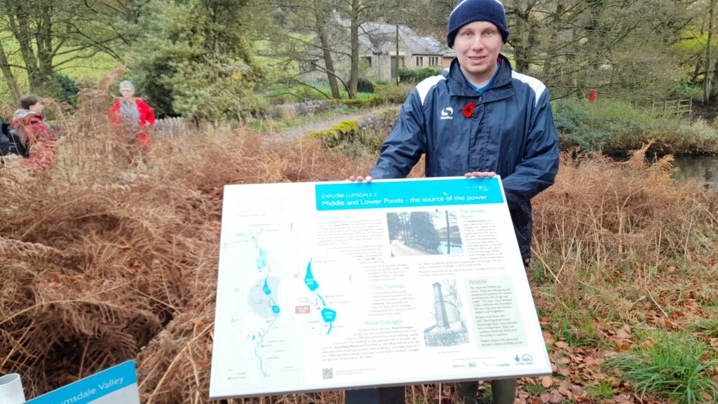 Walker standing by an information board about local ponds – A walker stands next to an informational sign about the Middle and Lower Ponds, surrounded by tall ferns and trees.