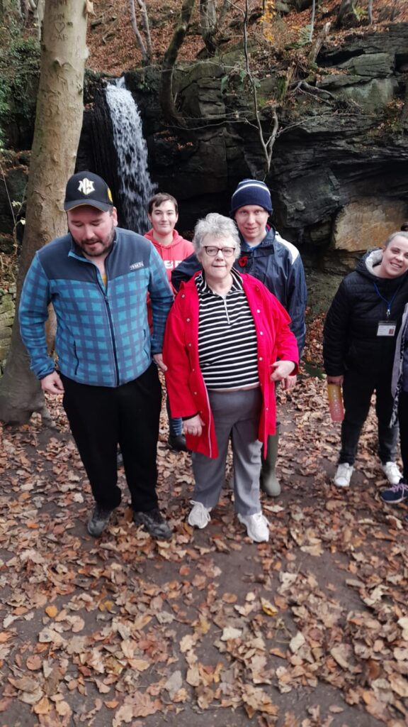 Group of walkers posing by a waterfall in the woods – A group of walkers standing together in front of a cascading waterfall in a forest area covered in autumn leaves.