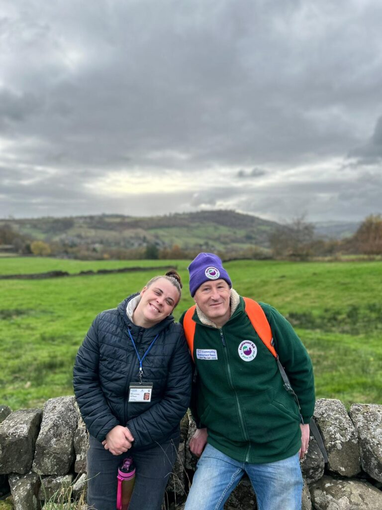 Two walkers in front of a stone wall with countryside views – Two walkers leaning against a stone wall, smiling with a green landscape and hills in the background.