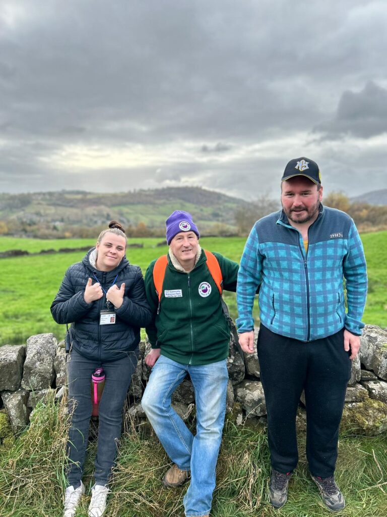 Three walkers by a stone wall with a rolling countryside view – Three walkers standing in front of a stone wall, with a scenic countryside and cloudy sky in the background.