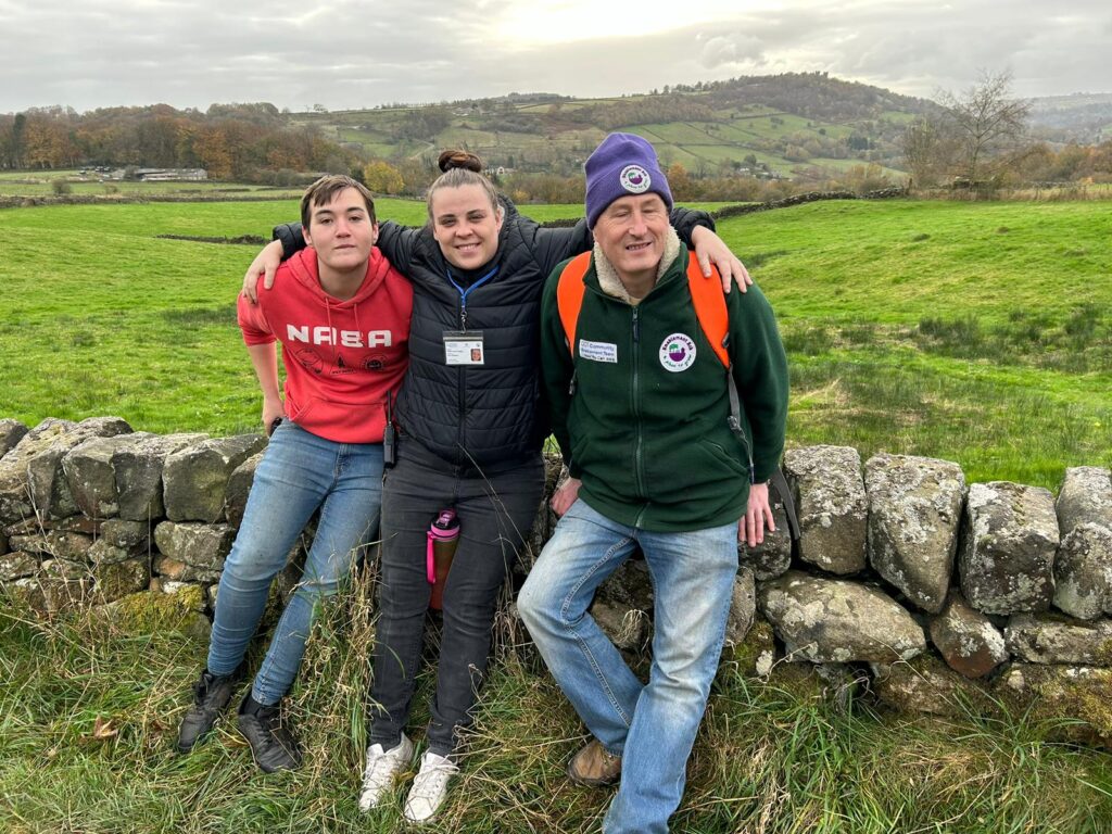 Three walkers posing by a stone wall with green fields behind – A group of three walkers standing together by a stone wall with expansive green fields and hills in the distance.