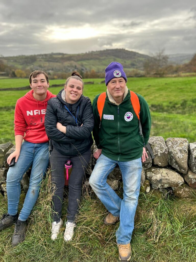 Three walkers sitting on a stone wall with a countryside backdrop – Three smiling walkers sit together on a stone wall, with rolling hills and green fields stretching into the background under a cloudy sky.