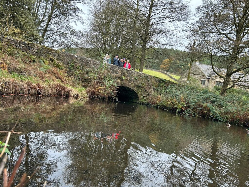 Group of walkers on a stone bridge over a reflective stream – A group of walkers standing on a stone bridge, looking down at the calm water that reflects the trees and landscape around them.