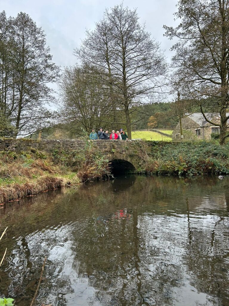 Walkers on a stone bridge with lush countryside around – A group of walkers gathered on a stone bridge over a stream, surrounded by tall trees and grassy hills.