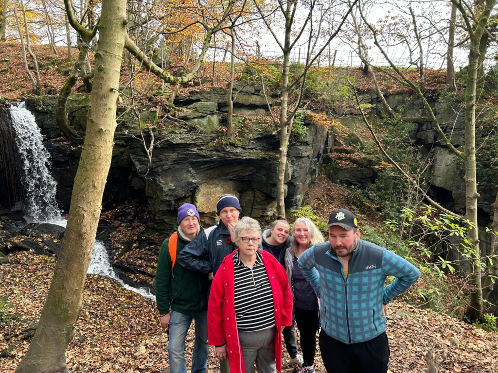 Group of walkers in front of a waterfall in a rocky forest area – A group of walkers pose in front of a cascading waterfall, surrounded by rocky terrain and autumn foliage in a forest setting.