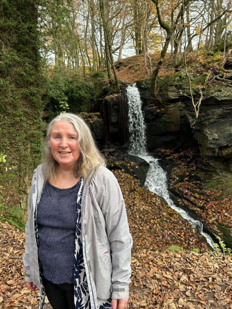 Smiling woman in front of a waterfall – A woman smiles at the camera, standing in front of a cascading waterfall with a background of autumn leaves and forest trees.