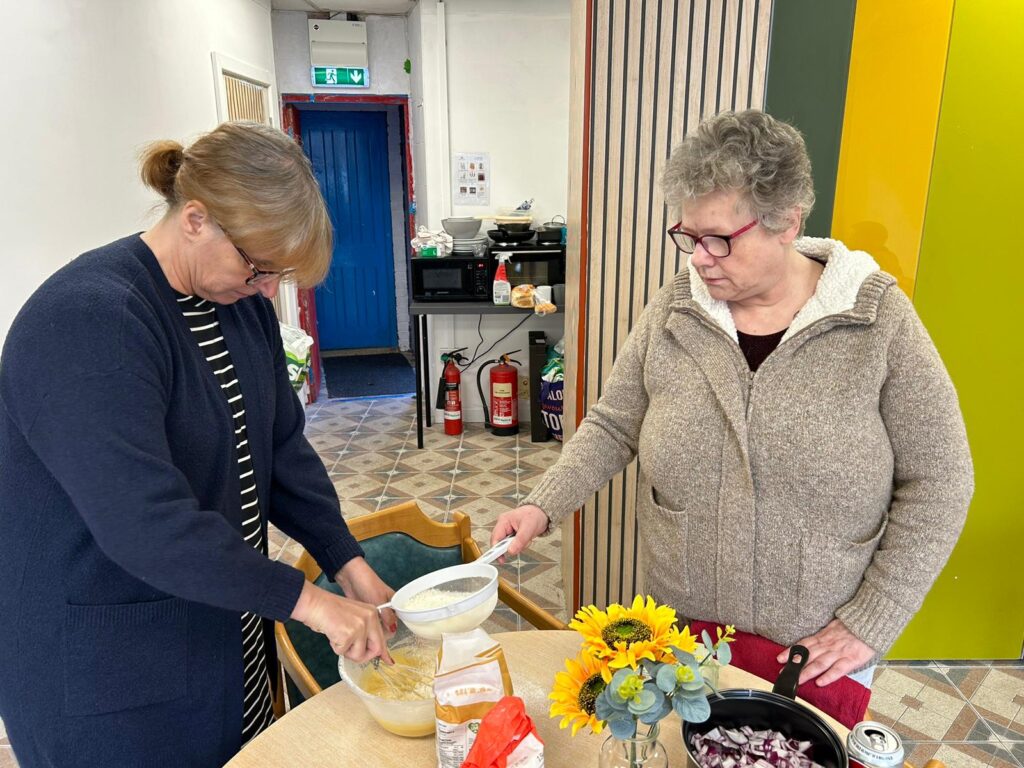 Two members of The Hub Cooking Club preparing ingredients at a table. One is sifting flour into a mixing bowl while the other looks on, both focused on their task. A small vase of sunflowers sits on the table, along with some chopped onions ready for cooking.