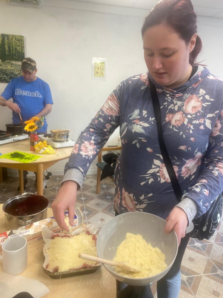 A member of The Hub Cooking Club preparing a berry crumble. She is holding a mixing bowl filled with crumble topping, sprinkling it over the tray bake. In the background, another member is stirring a pot on a portable stove, with a small vase of sunflowers adding a cheerful touch to the kitchen workspace.