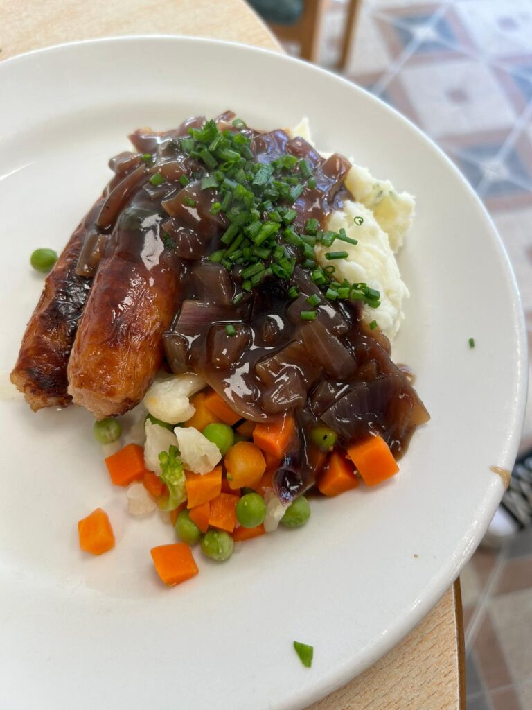 A plate of bangers and mash served with mixed vegetables and rich onion gravy, garnished with freshly chopped chives. The sausages are golden brown, and the mashed potatoes are creamy, making for a classic, comforting meal.