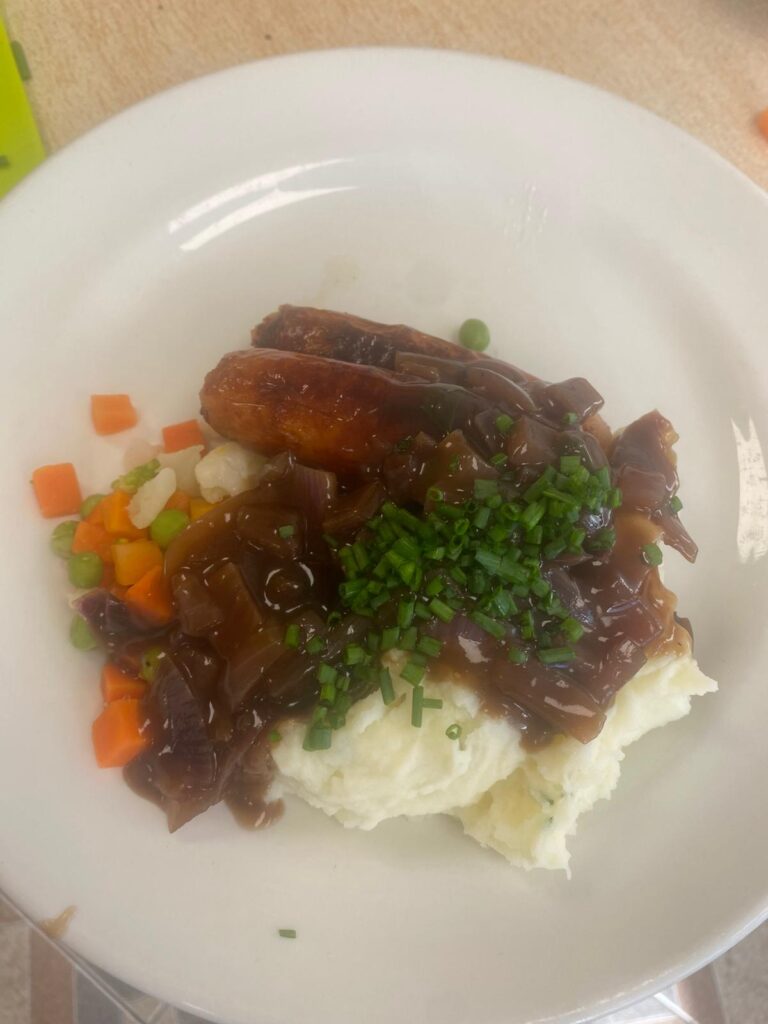 A close-up of a serving of bangers and mash, featuring two sausages topped with rich onion gravy, creamy mashed potatoes, and a garnish of fresh chives. Mixed vegetables are placed alongside, adding a pop of color to this comforting meal.