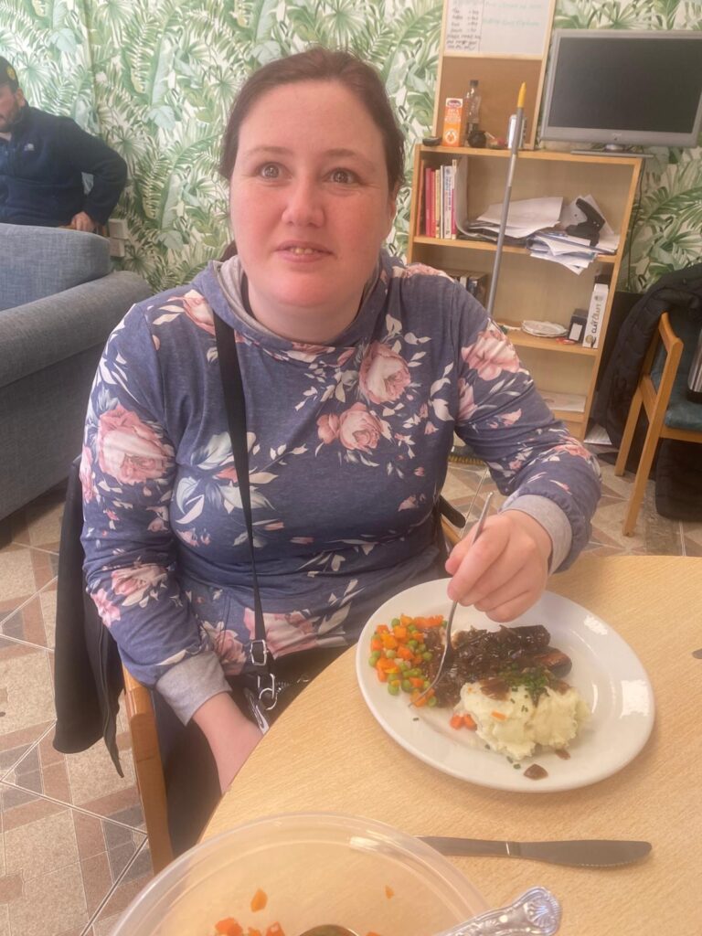 A member of The Hub Cooking Club sitting at a table, smiling and enjoying a plate of bangers and mash with mixed vegetables. She is holding a fork, ready to dig in. The room has a bright leaf-patterned wallpaper, adding a cheerful backdrop to the scene.