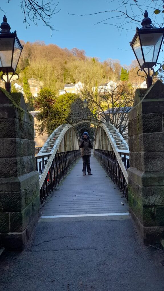 A CET Walking Club member standing at the centre of Jubilee Bridge in Matlock, surrounded by scenic winter trees and historic streetlamps, waving joyfully.