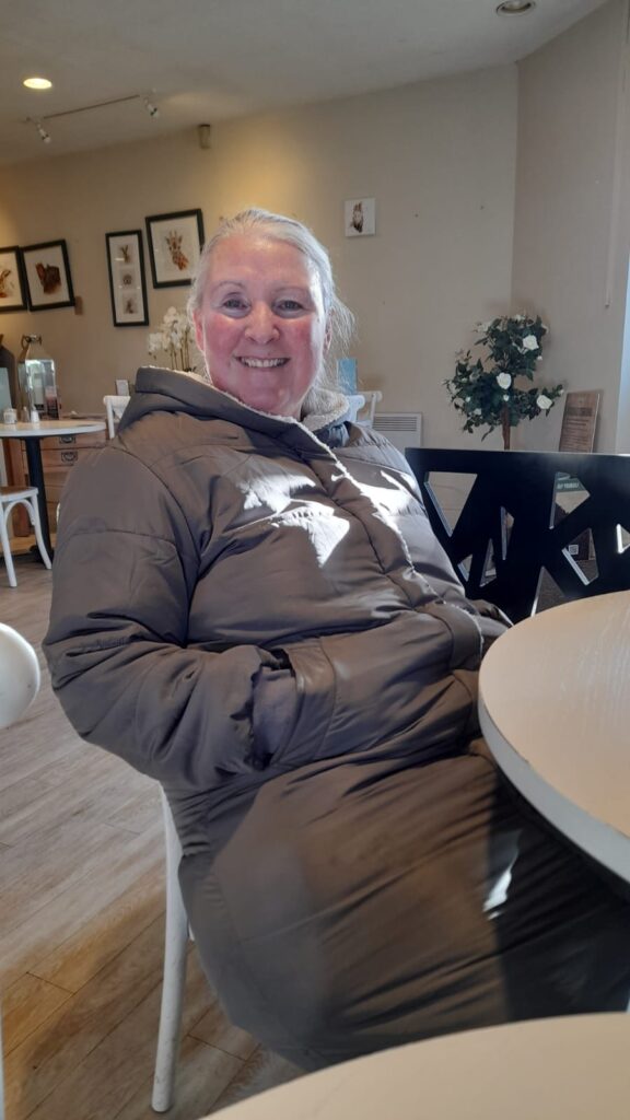 A cheerful CET Walking Club member seated inside the cosy café at Matlock Garden Centre, wearing a warm winter coat and smiling warmly at the camera.