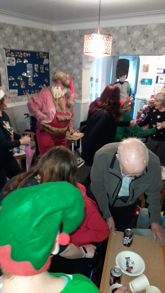 Santa Claus interacting with attendees at a lively Christmas celebration, with festive decorations, including a bulletin board and holiday posters, in the background.