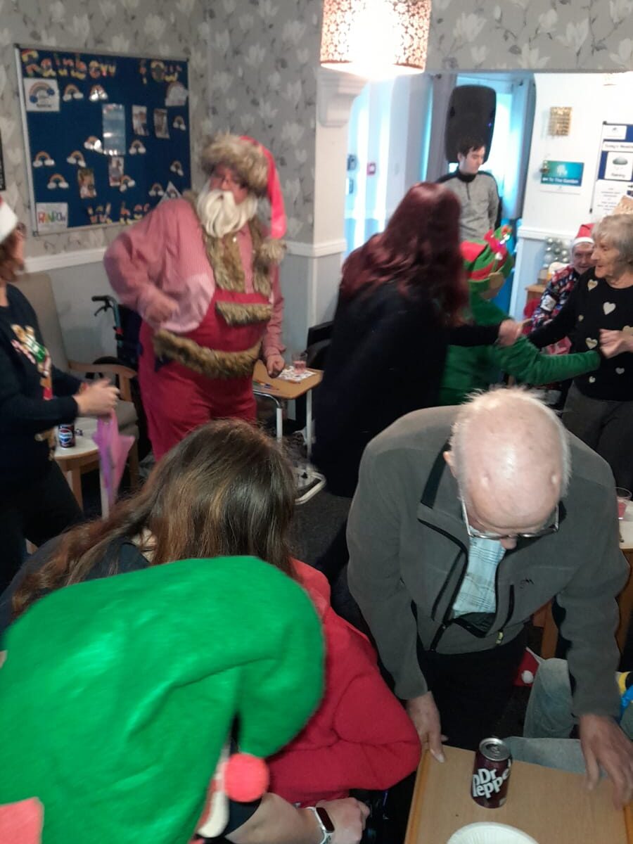 Santa Claus interacting with attendees at a lively Christmas celebration, with festive decorations, including a bulletin board and holiday posters, in the background.