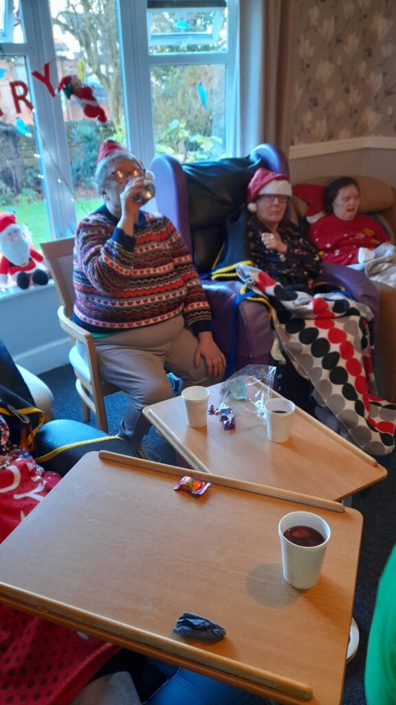 A group of people sitting in a cosy room near a large window decorated with festive ornaments. One person in a colourful Christmas jumper and Santa hat is sipping from a can, while others, wrapped in blankets, enjoy the holiday atmosphere. Tables in front of them hold cups, sweets, and small snacks.