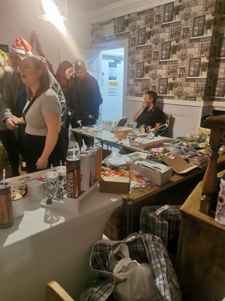 A bustling room with Christmas stalls and visitors browsing items. A woman in a Santa hat stands in the foreground, while others are engaged in conversation and shopping.