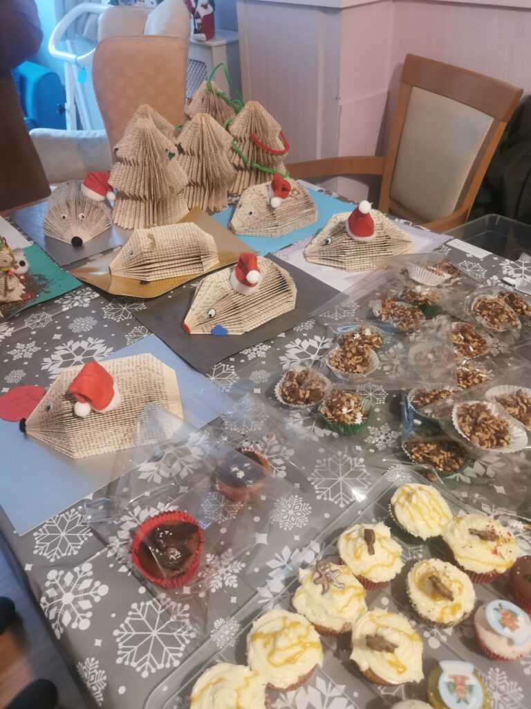 A table featuring handmade crafts, including folded book Christmas trees and hedgehogs with Santa hats, alongside a variety of festive cupcakes and treats.