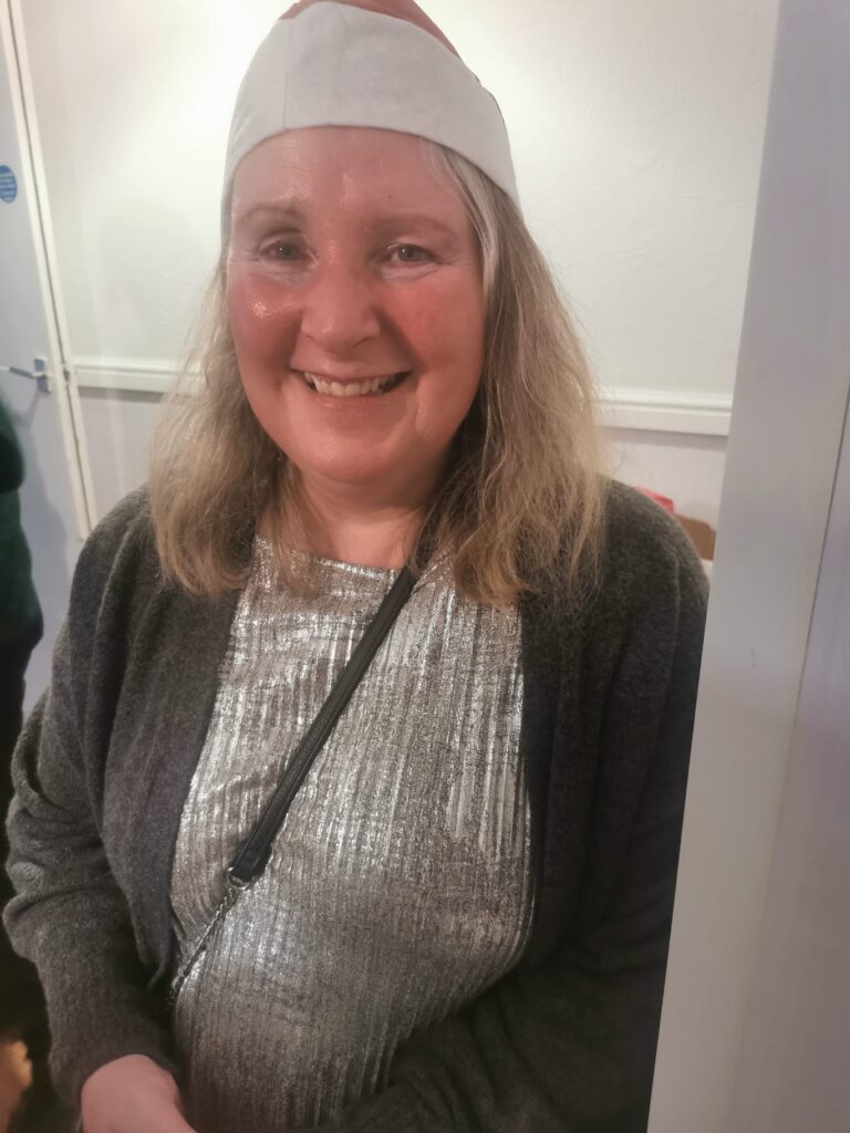 A smiling woman wearing a Santa hat and a shimmering silver top, posing for the camera in a festive setting.