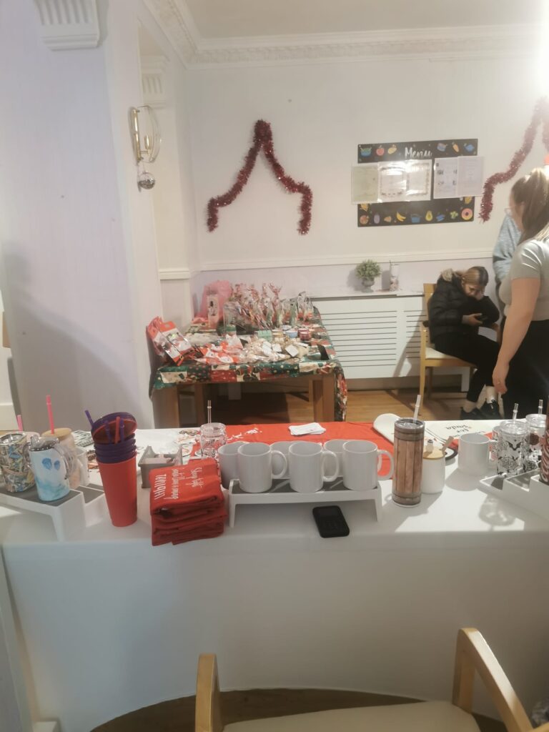 A display table with festive mugs, tumblers, and gift items, surrounded by Christmas decorations, including red tinsel hanging on the wall.