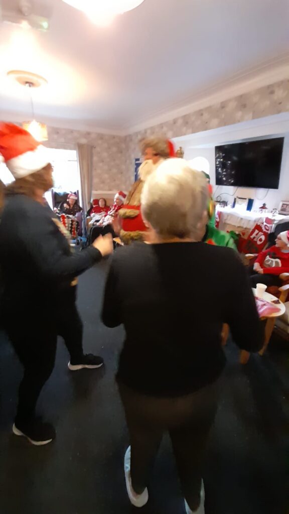 Santa Claus and elves entertaining a group of people in a festive room filled with holiday decorations and a television in the background.