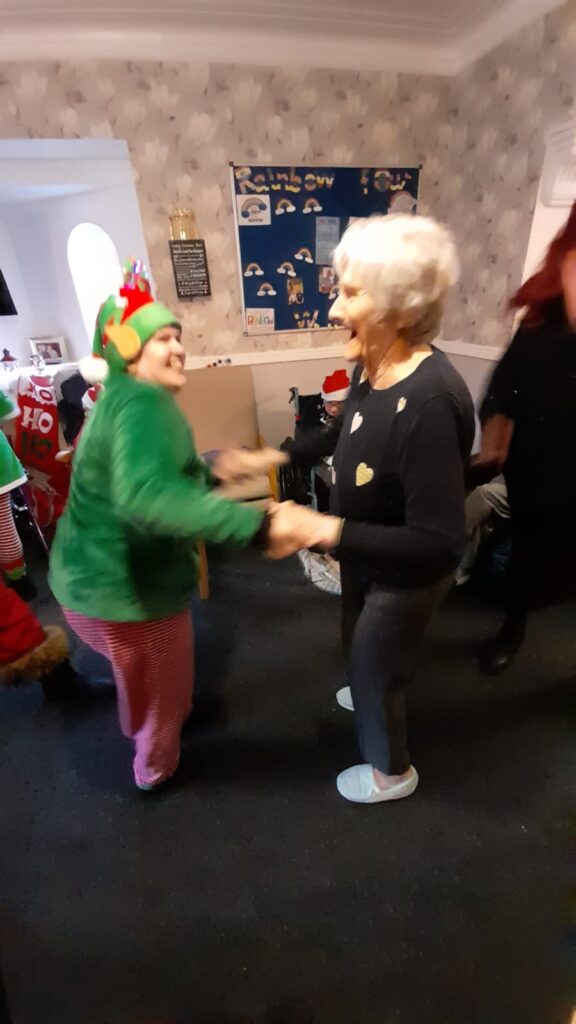 An elf in a green costume dancing with a smiling older woman in a cosy room decorated with holiday-themed crafts and posters.