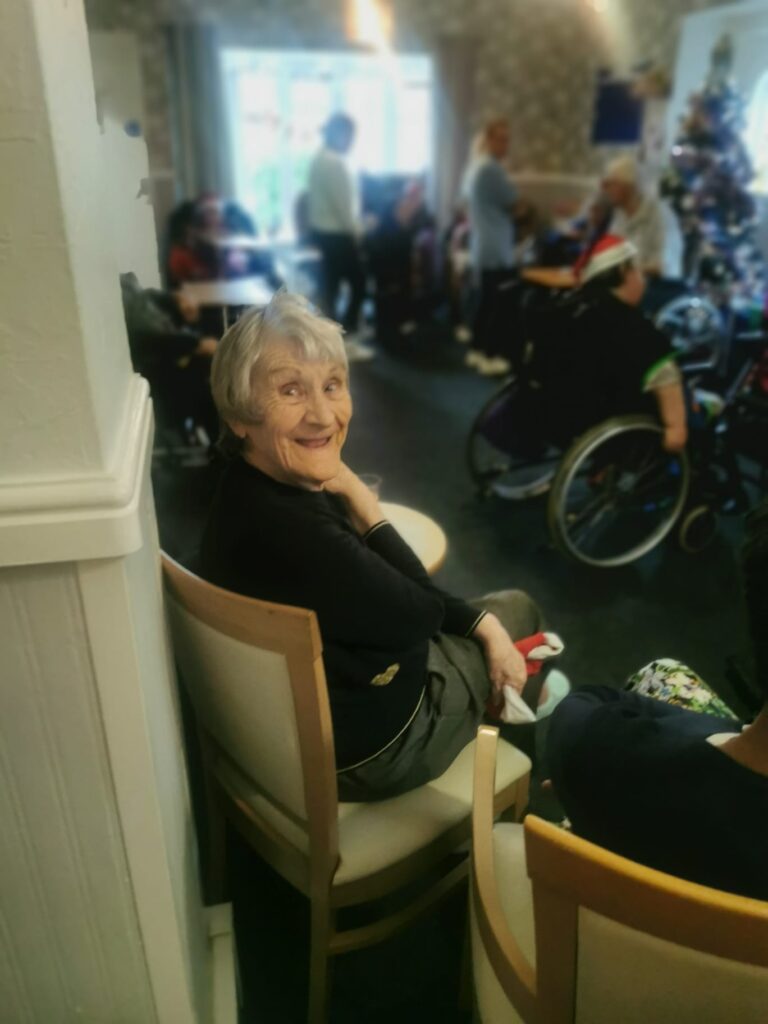 A cheerful elderly woman sitting in a chair, smiling at the camera while others in the room enjoy a festive gathering.