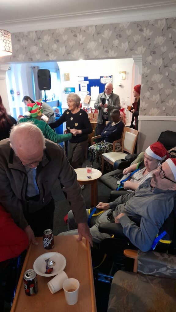 A room filled with people engaged in a holiday party, with some seated and others dancing. Festive hats, drinks, and snacks are visible on the tables.