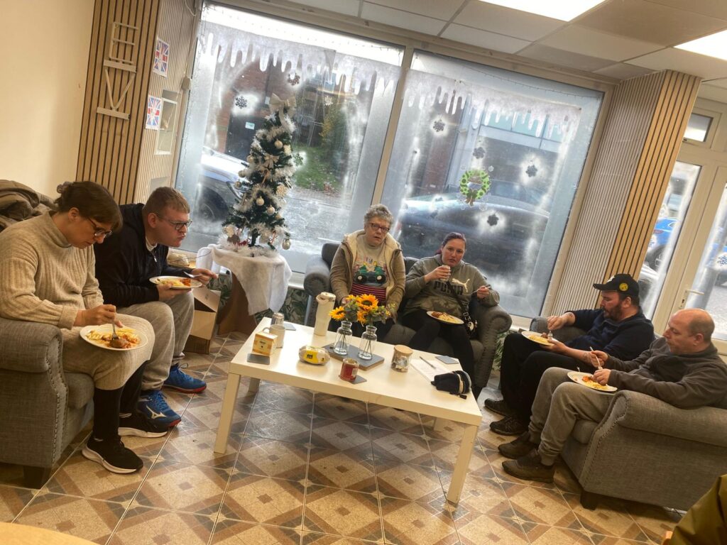  A similar scene to the previous image, showing a group of people gathered in a festive room with Christmas decorations and enjoying their meals together.