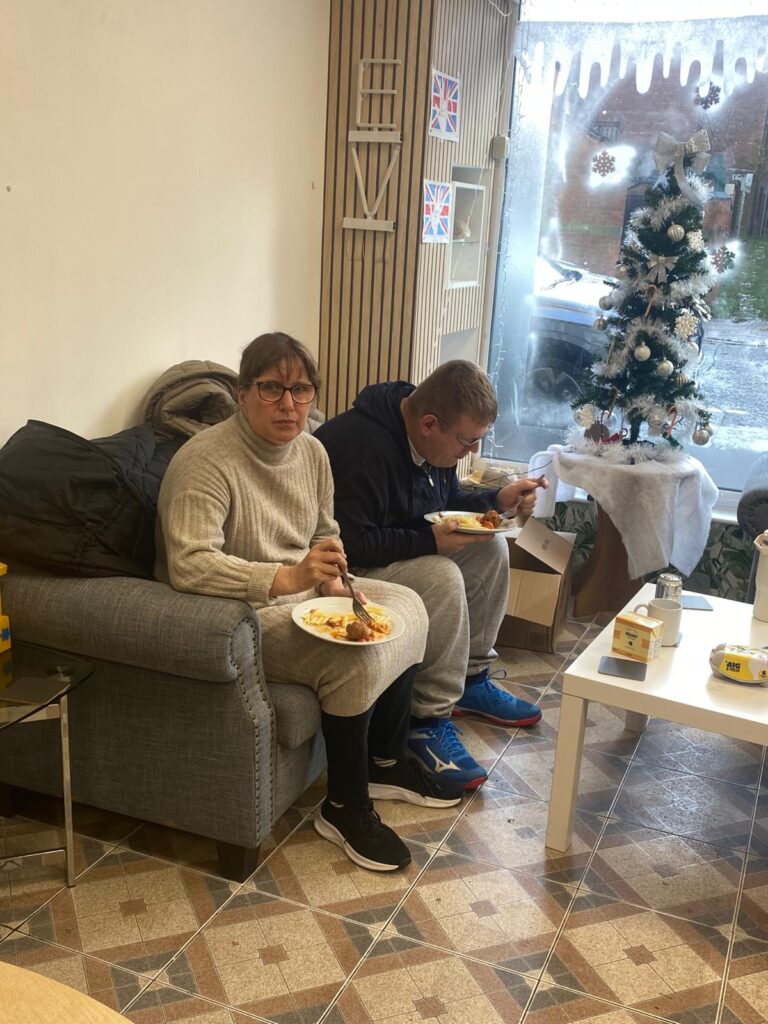 Two individuals sitting on a couch enjoying their plates of food in a warmly decorated room. A small Christmas tree and winter-themed window decorations add to the festive atmosphere.