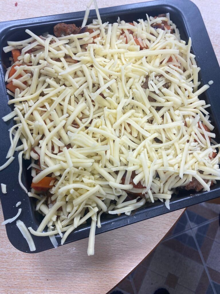 A casserole dish filled with a hearty mix of vegetables, meatballs, and tomato sauce, topped generously with grated cheese, ready for baking.