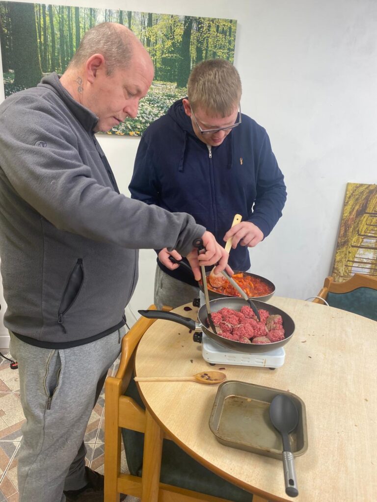 Two individuals working together to cook. One is flipping meatballs in a pan while the other stirs tomato sauce. The cooking takes place on a round table, and a peaceful forest scene decorates the wall.