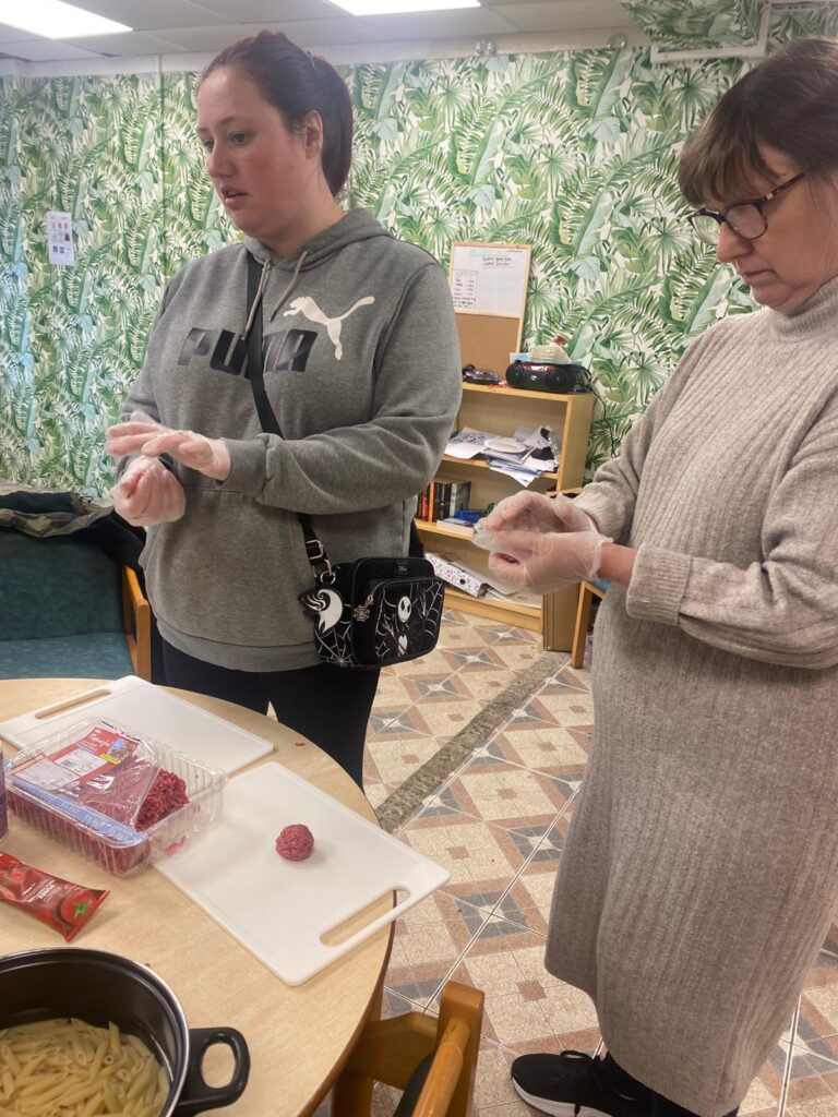 Two women shaping meatballs on cutting boards at a table. Ingredients such as ground beef and pasta are visible nearby, and the background features a green leafy wallpaper.