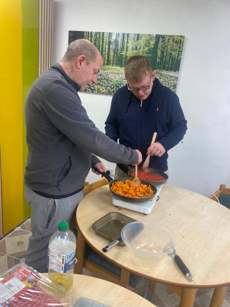 Two individuals cooking together at a table. One person stirs a pan of sautéed vegetables, while the other prepares a pan of tomato sauce. A serene forest-themed wall decoration is visible in the background.