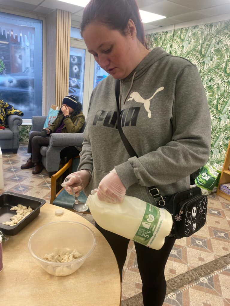 A woman carefully pouring milk into a mixing bowl while preparing a dish. She wears gloves for hygiene, and a festive room with winter decorations is visible in the background.