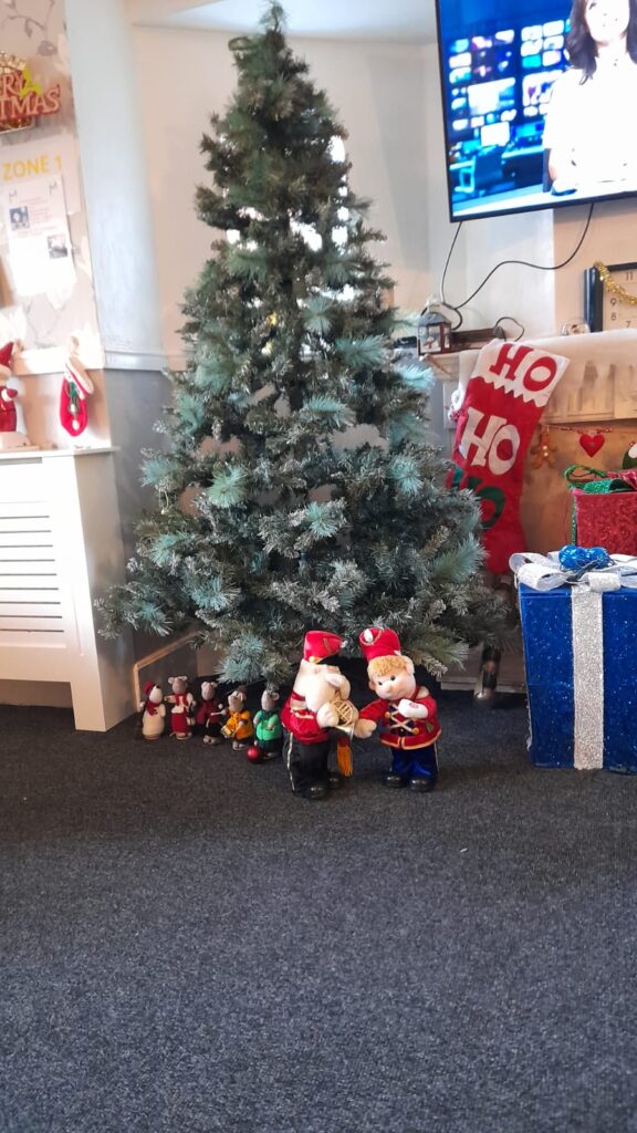 A Christmas tree surrounded by small festive decorations, including toy soldiers, wrapped presents, and a line of knitted mice in holiday costumes. A large stocking with "HO HO HO" hangs nearby, adding to the festive atmosphere.