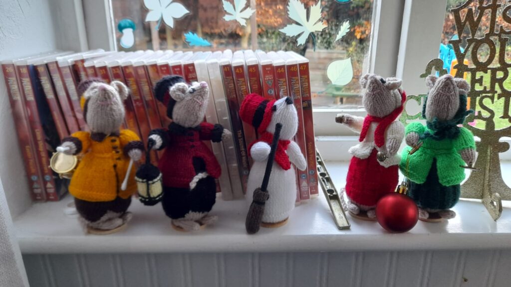 A windowsill display featuring an adorable set of hand-knitted mice dressed in festive outfits, including scarves, lanterns, and hats, set against a background of DVDs and winter-themed window stickers.