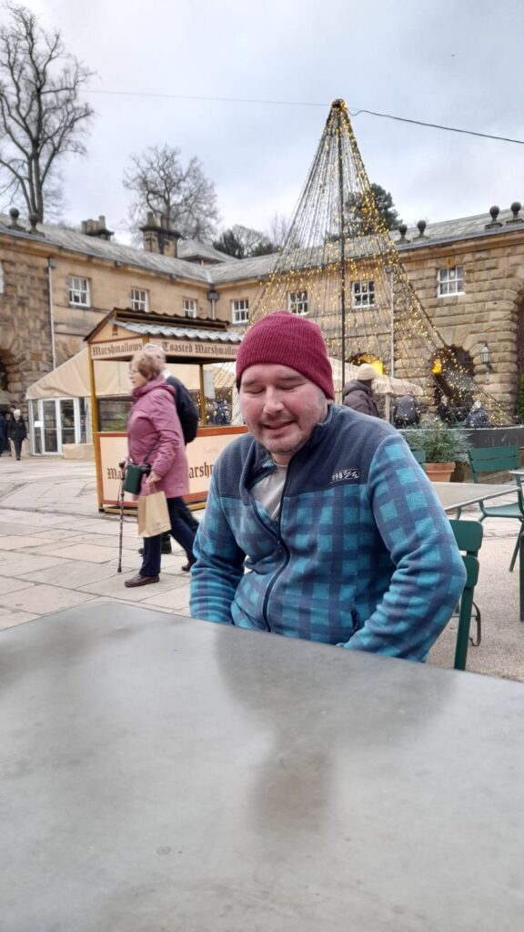 A man in a red beanie and blue checkered jacket sits at an outdoor café table, smiling with his eyes closed. Behind him, a festive wire Christmas tree lit with golden lights stands in front of a stone building. Other people are seen walking around the market area.