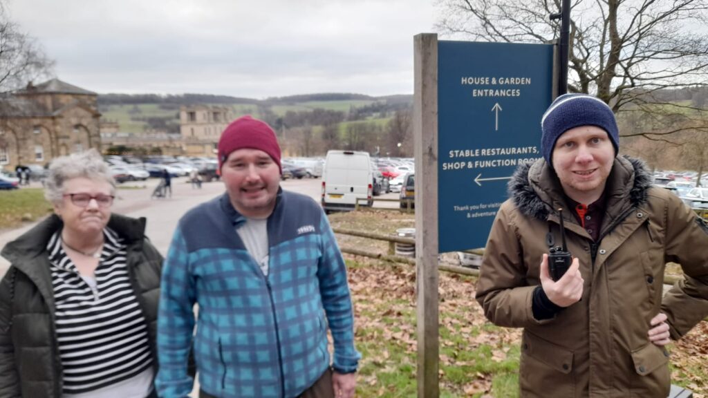 Three people stand near the "House & Garden Entrances" sign at Chatsworth House. From left to right: an older woman in a green coat and glasses, a man in a red beanie and blue checkered jacket, and a man in a brown coat holding a small device. Behind them are parked cars, a stone building, and rolling hills.