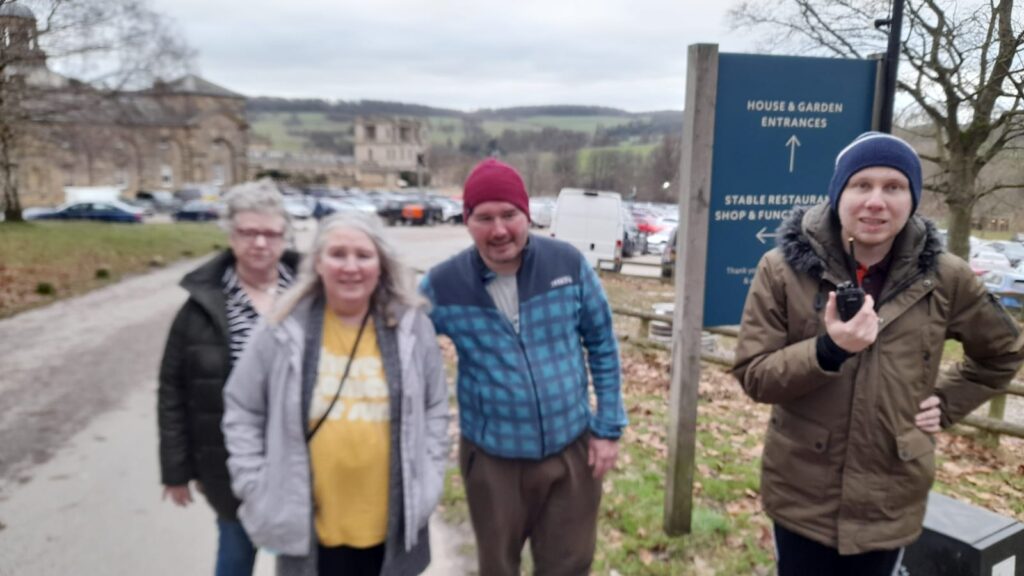 A group of four people pose near a sign reading "House & Garden Entrances" at Chatsworth House. The group consists of an older woman in a green coat, a woman in a grey coat and yellow t-shirt, a man in a red beanie and blue jacket, and another man in a brown coat holding a small device. The background features parked cars and the rolling hills of Chatsworth.