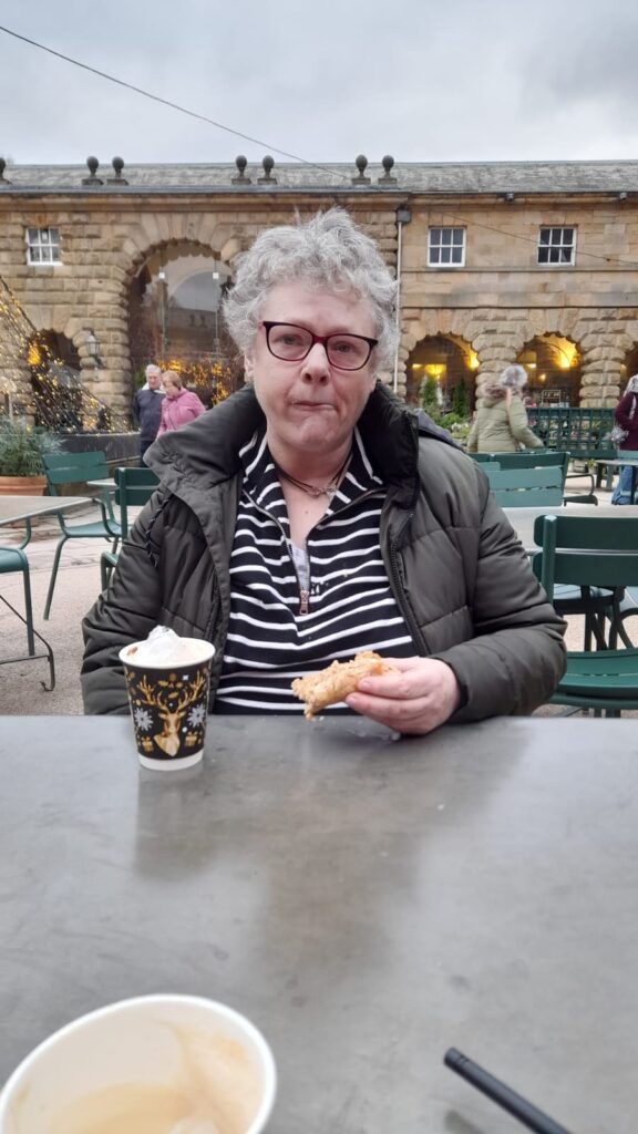 An older woman with glasses and short grey hair sits at an outdoor café table, holding a pastry in one hand. In front of her is a festive takeaway cup with a reindeer design. Behind her, stone arches are decorated with twinkling lights, and people can be seen walking through the area.